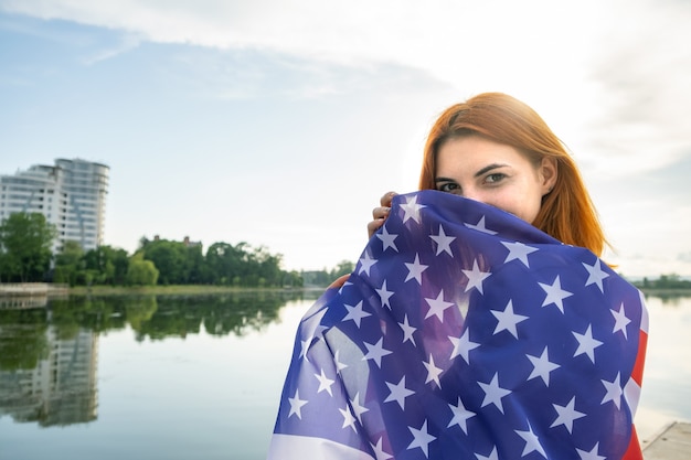 Szczęśliwa Rudowłosa Dziewczyna Z Flagą Narodową Usa Na Ramionach. Pozytywna Młoda Kobieta Z Okazji Dnia Niepodległości Stanów Zjednoczonych.