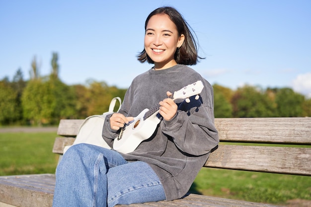 Szczęśliwa śliczna dziewczyna siedzi samotnie na ławce w parku, gra na gitarze ukulele i cieszy się słonecznym dniem na świeżym powietrzu