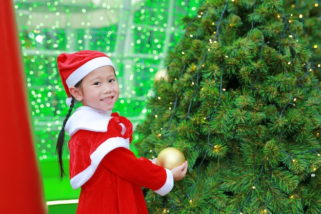Szczęśliwa śliczna mała Azjatycka dziecko dziewczyna w Santa kostiumowej pobliskiej choince i tle.