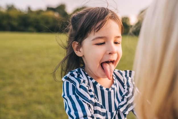 Szczęśliwa śliczna mała dziewczynka bawi się z jej ładną matką pokazując język w parku Kochająca matka i córka spędzają razem czas w parku Macierzyństwo i dzieciństwo Koncepcja emocji rodzinnych ludzi
