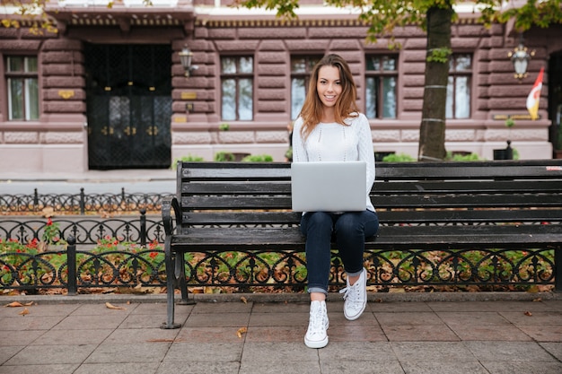 Szczęśliwa śliczna młoda kobieta siedzi na ławce i korzysta z laptopa na zewnątrz