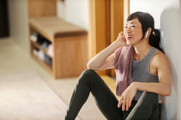 Szczęśliwa, sprawna japońska kobieta siedzi w domu na podłodze i cieszy się muzyką po treningu.