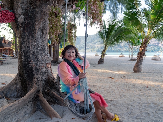 szczęśliwa starsza azjatycka kobieta w kolorowej sukience usiądź i zrelaksuj się na huśtawce pod wielkim drzewem na nadmorskiej plaży