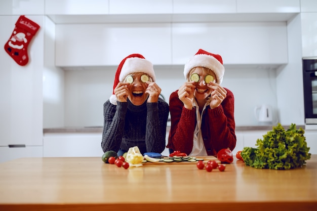 Szczęśliwa starsza kobieta i jej córka, opierając się na kuchennym blacie i trzymając plasterki ogórka na oczach. Obaj mają na głowach czapki Mikołaja. Na blacie kuchennym są warzywa.