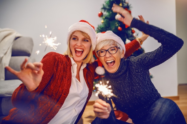 Szczęśliwa starsza kobieta i jej córka świętuje nowy rok. Obie mają na głowach czapki Mikołaja i trzymają zimne ognie. W tle jest choinka. Pojęcie wartości rodzinnych.
