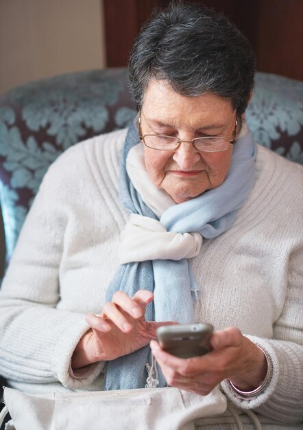Szczęśliwa starsza kobieta, korzystająca ze smartfona, który wysyła SMS-y, przeglądając telefon komórkowy, wysyłając wiadomości, siedząc na kanapie w domu