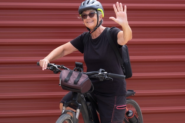 Szczęśliwa starsza kobieta rowerzysta na wycieczce na świeżym powietrzu w pobliżu czerwonego metalowego panelu cieszącego się zdrowym stylem życia