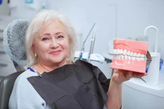 Szczęśliwa starsza kobieta uśmiecha się do kamery, trzymając model dentystyczny, siedząc w gabinecie dentystów