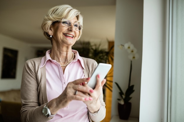 Szczęśliwa starsza kobieta wiadomości tekstowe na telefonie komórkowym, patrząc przez okno w domu