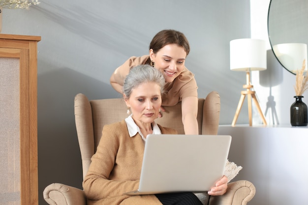 Szczęśliwa starsza matka środkowego siedzi na krześle z córką, patrząc na laptopa. Młoda kobieta pokazuje wideo, zdjęcia mamusi, zaufane relacje. Koncepcja rodziny.