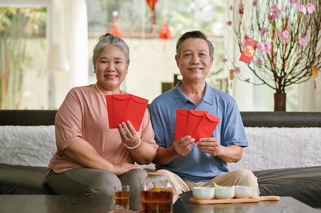 Szczęśliwa starsza para siedzi na kanapie w domu i pokazuje czerwone koperty przygotowane na tet
