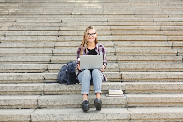 Szczęśliwa studentka dziewczyna siedzi na schodach, pracując z laptopem, przygotowując się do egzaminów i pijąc kawę na świeżym powietrzu, w kampusie uniwersyteckim. Koncepcja technologii, edukacji i pracy zdalnej, kopia przestrzeń