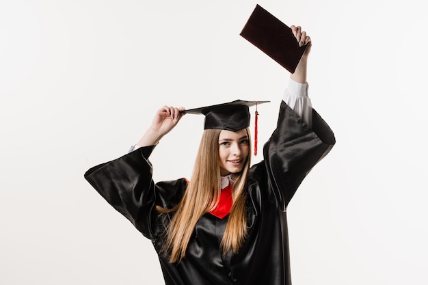 Szczęśliwa studentka w czarnej sukni dyplomowej i czapce podnosi dyplom magistra nad głową na białym tle Absolwentka kończy studia i świętuje osiągnięcia akademickie