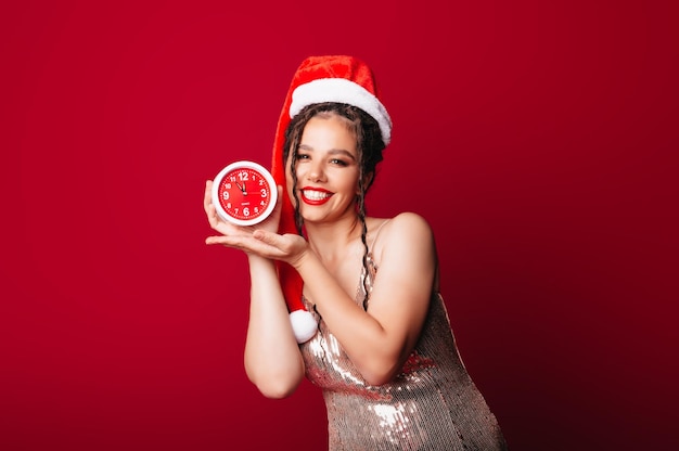 Szczęśliwa urocza kobieta w kapeluszu Świętego Mikołaja trzyma budzik na czerwonym tle Pojęcie sezonu Bożego Narodzenia Boże Narodzenie i nowy rok