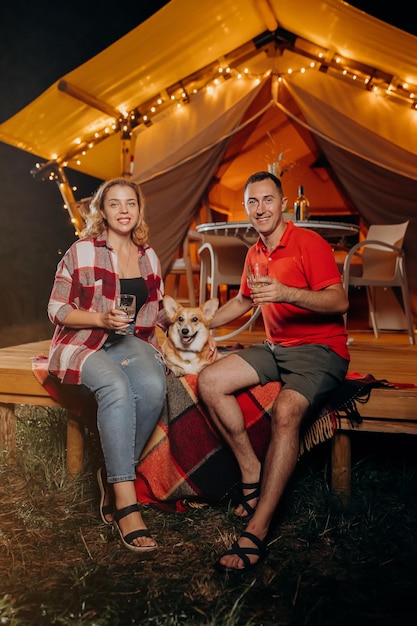 Szczęśliwa urocza para z psem Welsh Corgi Pembroke relaksująca się wieczorem w glampingu i pijąca wino w pobliżu przytulnego ogniska Luksusowy namiot kempingowy do rekreacji na świeżym powietrzu i rekreacji Koncepcja stylu życia
