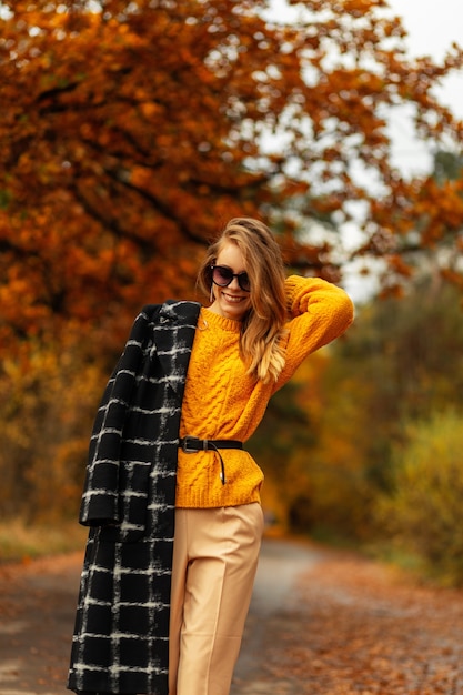 Szczęśliwa Uśmiechnięta Dziewczyna W Modnych Jesiennych Ubraniach Z żółtym Vintage Swetrem, Czarnym Płaszczem I Okularami Przeciwsłonecznymi Spaceruje Po Parku Z Kolorowymi Jesiennymi Liśćmi