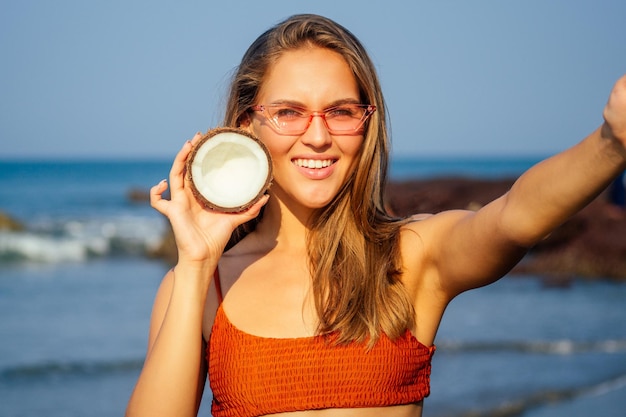 Szczęśliwa uśmiechnięta dziewczyna w stylowym pomarańczowym koralowym stroju kąpielowym biorąca selfie na telefon z kokosem w vacationbikini kobieta trzyma telefon komórkowy zdjęcie aparat smartfon zdjęcia na oceanie indiaspa coco nut oil