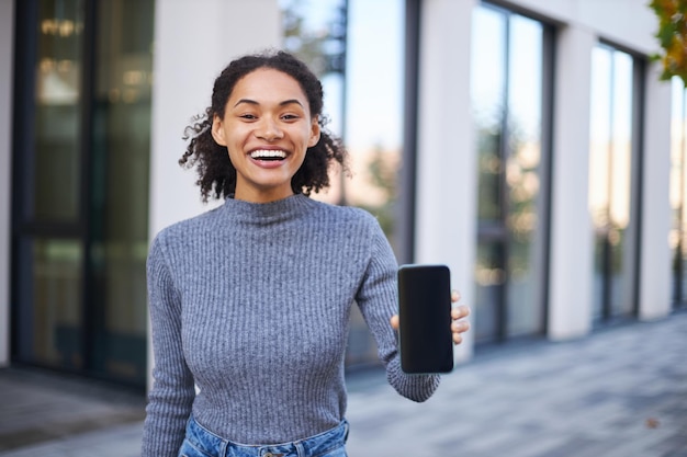 Szczęśliwa uśmiechnięta etniczna młoda kobieta pokazująca pusty wyświetlacz nowoczesnego smartfona na ulicy miasta