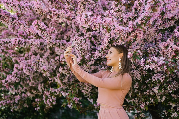 Szczęśliwa uśmiechnięta kobieta robi selfie na swoim smartfonie w kwitnącym wiosną ogrodzie, ciesząc się przyrodą