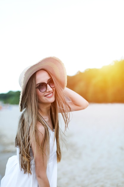 Szczęśliwa uśmiechnięta kobieta w błogości wolnego szczęścia na plaży oceanu stojąca i pozująca z kapeluszem i okularami przeciwsłonecznymi. Portret modelki w białej letniej sukience cieszącej się przyrodą.