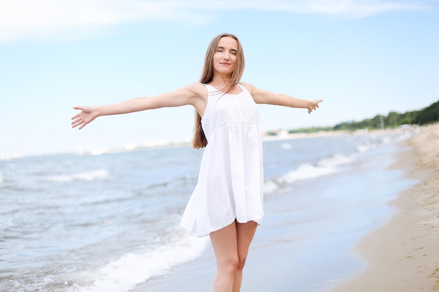 Szczęśliwa uśmiechnięta kobieta w błogości wolnego szczęścia na plaży oceanu stojąca z otwartymi rękami. Portret wielokulturowej modelki w białej letniej sukience cieszącej się przyrodą podczas wakacji wakacyjnych