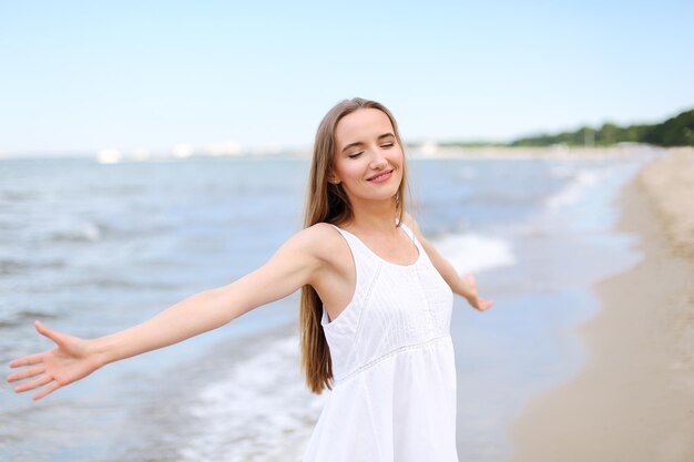 Szczęśliwa uśmiechnięta kobieta w błogości wolnego szczęścia na plaży oceanu stojąca z otwartymi rękami. Portret wielokulturowej modelki w białej letniej sukience cieszącej się przyrodą podczas wakacji wakacyjnych