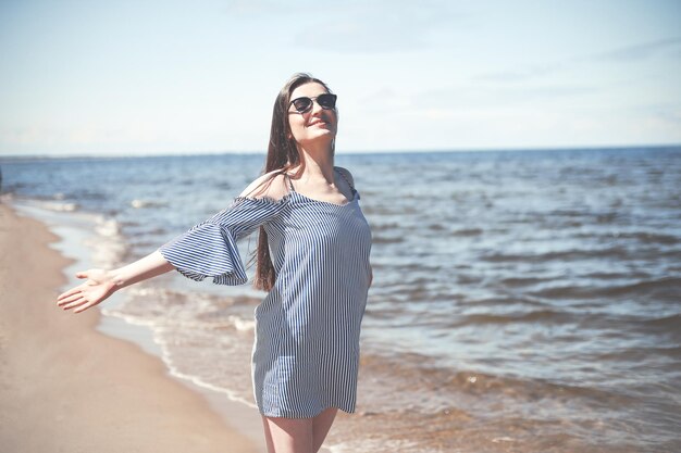 Szczęśliwa uśmiechnięta kobieta w wolnej błogości na plaży oceanu stojąca z otwartymi rękami. Portret brunetki modelki w letniej sukience cieszącej się przyrodą podczas wakacji na świeżym powietrzu.