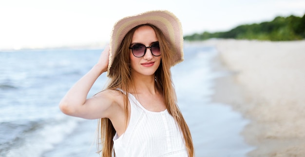 Szczęśliwa uśmiechnięta kobieta w wolnym szczęściu na plaży oceanu stojąca i pozująca z kapeluszem i okularami przeciwsłonecznymi. Portret kobiety w białej letniej sukience cieszącej się naturą.