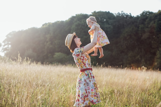 Szczęśliwa uśmiechnięta ładna matka w sukience i słomkowym kapeluszu, trzymająca i wychowująca małą córeczkę bawiącą się w pięknym letnim polu