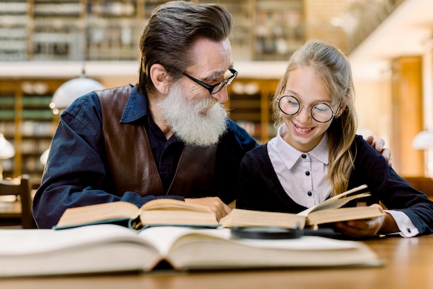 Szczęśliwa uśmiechnięta mała dziewczynka z jej rozochoconymi dziadek czytelniczymi książkami przy biblioteką. Uśmiechnięta mała dziewczynka z jej starszym nauczycielem studiuje wpólnie w bibliotece