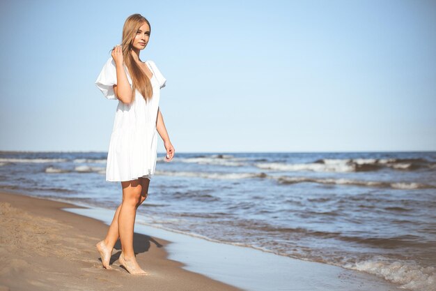 Szczęśliwa uśmiechnięta piękna kobieta chodzi po plaży oceanu w białej letniej sukience.
