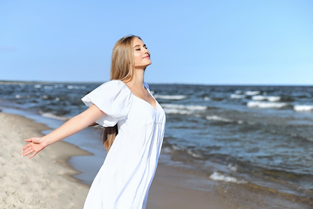 Szczęśliwa uśmiechnięta piękna kobieta jest na plaży oceanu w białej letniej sukience, otwarte ramiona