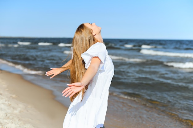 Szczęśliwa uśmiechnięta piękna kobieta jest na plaży oceanu w białej letniej sukience, z otwartymi ramionami.