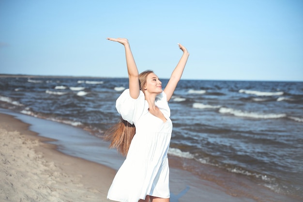 Szczęśliwa uśmiechnięta piękna kobieta na plaży oceanu, stojąca w białej letniej sukience, podnosząc ręce.