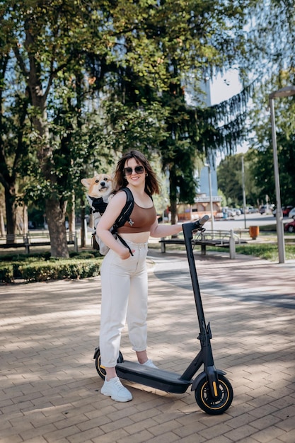 Szczęśliwa uśmiechnięta podróżniczka jedzie na swoim skuterze elektrycznym w miejskim parku z psem Welsh Corgi Pembroke w specjalnym plecaku