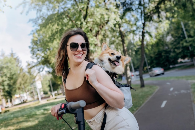 Szczęśliwa uśmiechnięta podróżniczka jedzie na swoim skuterze elektrycznym w miejskim parku z psem Welsh Corgi Pembroke w specjalnym plecaku