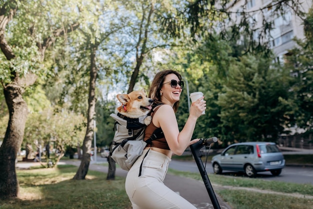 Szczęśliwa uśmiechnięta podróżniczka stoi z elektro skuterem i pije kawę w parku miejskim z psem Welsh Corgi Pembroke w specjalnym plecaku