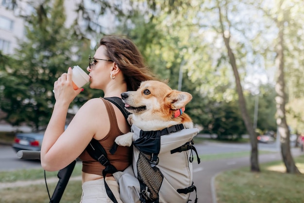 Szczęśliwa uśmiechnięta podróżniczka stoi z elektro skuterem i pije kawę w parku miejskim z psem Welsh Corgi Pembroke w specjalnym plecaku