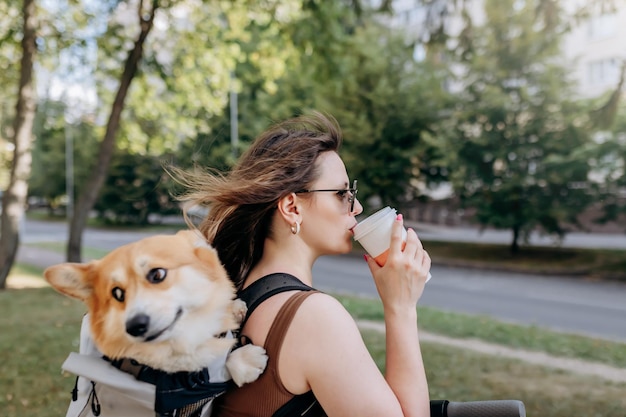 Szczęśliwa Uśmiechnięta Podróżniczka Stoi Z Elektro Skuterem I Pije Kawę W Parku Miejskim Z Psem Welsh Corgi Pembroke W Specjalnym Plecaku