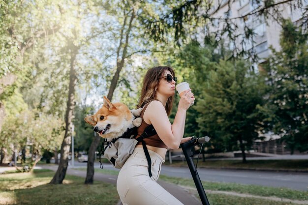 Szczęśliwa Uśmiechnięta Podróżniczka Stoi Z Elektro Skuterem I Pije Kawę W Parku Miejskim Z Psem Welsh Corgi Pembroke W Specjalnym Plecaku