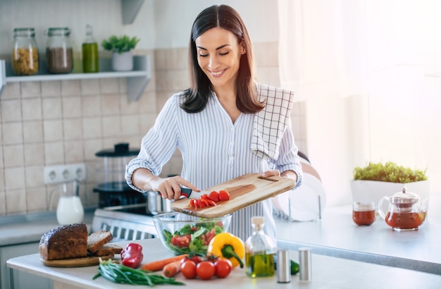 Szczęśliwa uśmiechnięta śliczna kobieta przygotowuje świeżą, zdrową wegańską sałatkę z wieloma warzywami w kuchni w domu i próbuje nowego przepisu