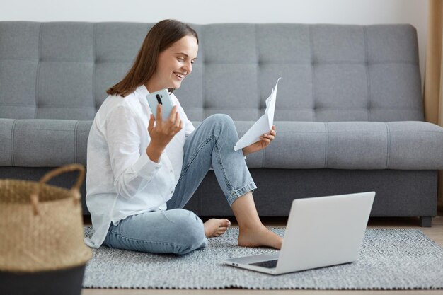 Szczęśliwa uśmiechnięta zadowolona kobieta ubrana w białą koszulę i dżinsy, siedząca na podłodze w pobliżu sofy, korzystająca z laptopa i telefonu z pozytywnym wyrazem twarzy, pracująca online w domu