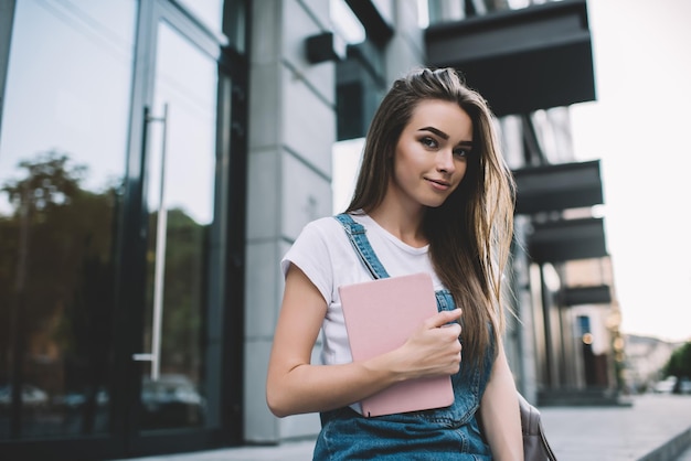 Szczęśliwa wesoła hipsterka w dżinsach uśmiechająca się podczas wolnego czasu w miejskim otoczeniu pozytywnej rasy kaukaskiej