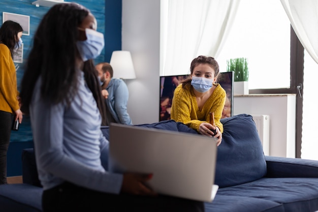 Szczęśliwa wesoła kobieta trzymająca butelkę piwa patrząca na laptopa rozmawiająca z afrykańskim przyjacielem w mieszkaniu w salonie nosząca maskę na twarz, aby zapobiec rozprzestrzenianiu się koronawirusa w czasie globalnej pandemii. Koncepcyjne i