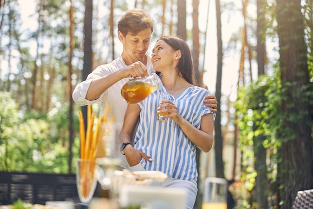 szczęśliwa wesoła para, podczas gdy mężczyzna nalewa pomarańczową lemoniadę do szklanki kobiety