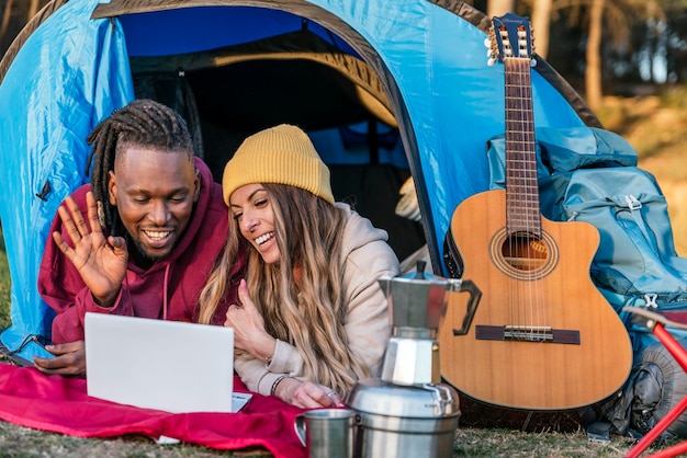 Szczęśliwa wielorasowa para leżąca w namiocie kempingowym za pomocą laptopa do rozmowy wideo z rodziną lub przyjaciółmi