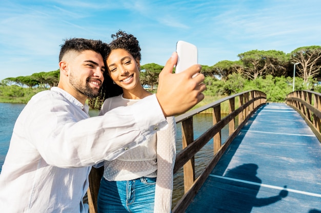 Zdjęcie szczęśliwa wielorasowa para zakochana pobyt na drewnianym moście przy selfie na wakacjach na łonie natury. przystojny facet fotografuje się z hiszpańską dziewczyną za pomocą smartfona. nowe nawyki technologiczne