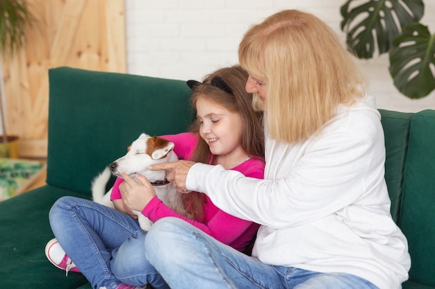 Szczęśliwa Wnuczka I Babcia Siedzi Na Kanapie Z Psem Jack Russell Terrier. Babcia Przytula Wnuka W Domu. Koncepcja Osób Starszych, Rodziny I Pokolenia.