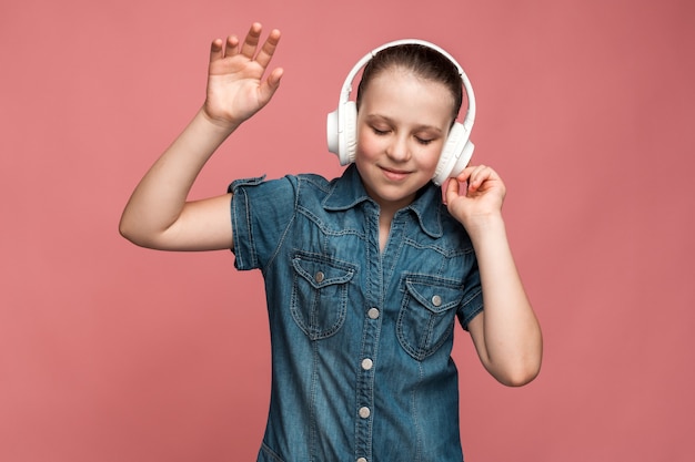 Szczęśliwa zabawna wielorasowa dziewczyna nosi bezprzewodowe słuchawki słuchania muzyki i czuje się dobrze z zadowoloną twarzą. Kryty strzał studio, na białym tle na różowym tle