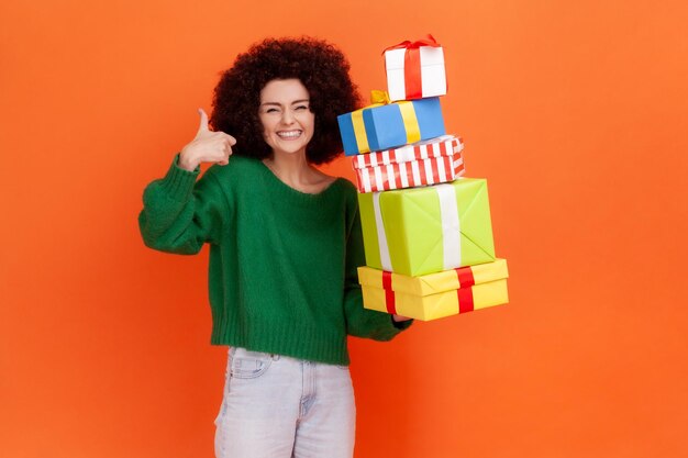 Szczęśliwa zadowolona kobieta z fryzurą Afro ubrana w zielony sweter w stylu casual stojący z dużą ilością obecnych pól, pokazując kciuk do góry, uśmiechając się. Kryty studio strzał na białym tle na pomarańczowym tle.